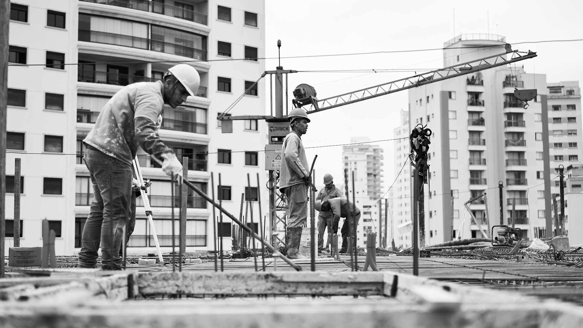 Operários montando armadura de laje em obra de edifício, com grua e prédios ao fundo